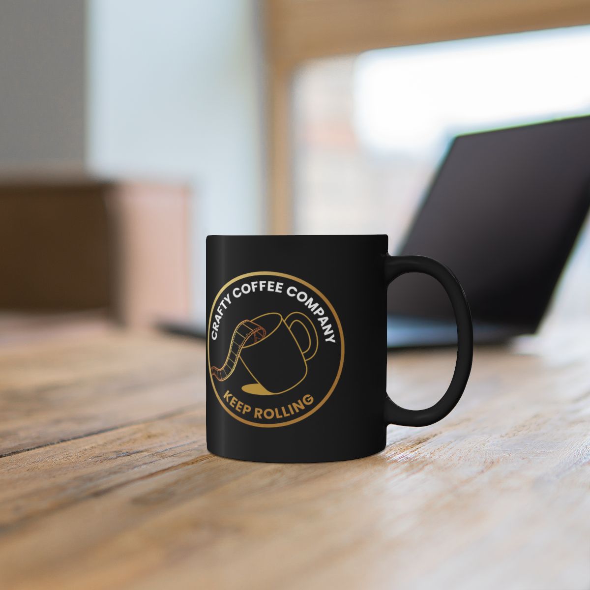 Black mug with the Crafty Coffee Company logo on a wooden surface in front of a laptop computer